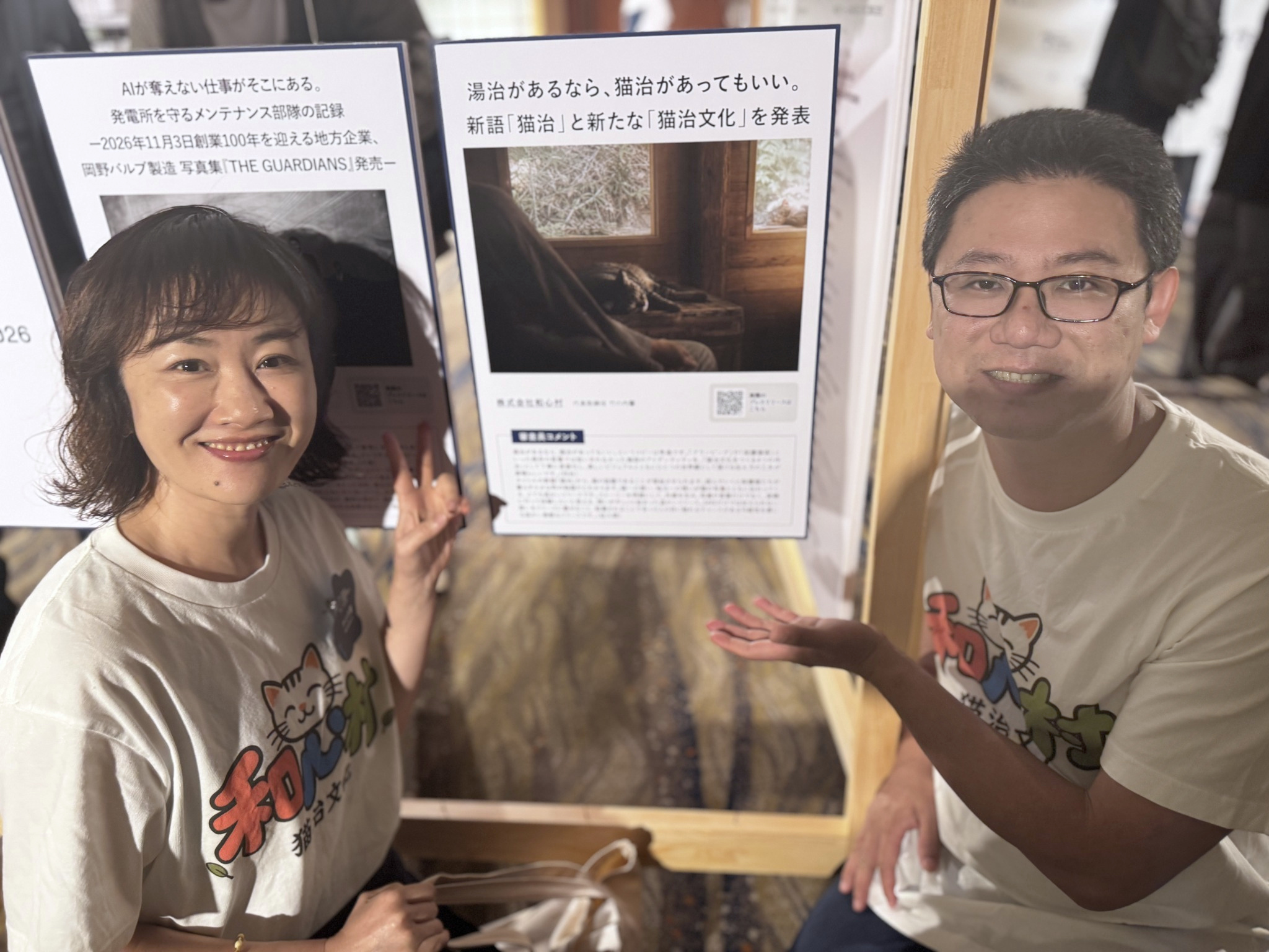 和心村・猫治文化のTシャツ姿の二人と、会場に展示された猫治文化の紹介パネル